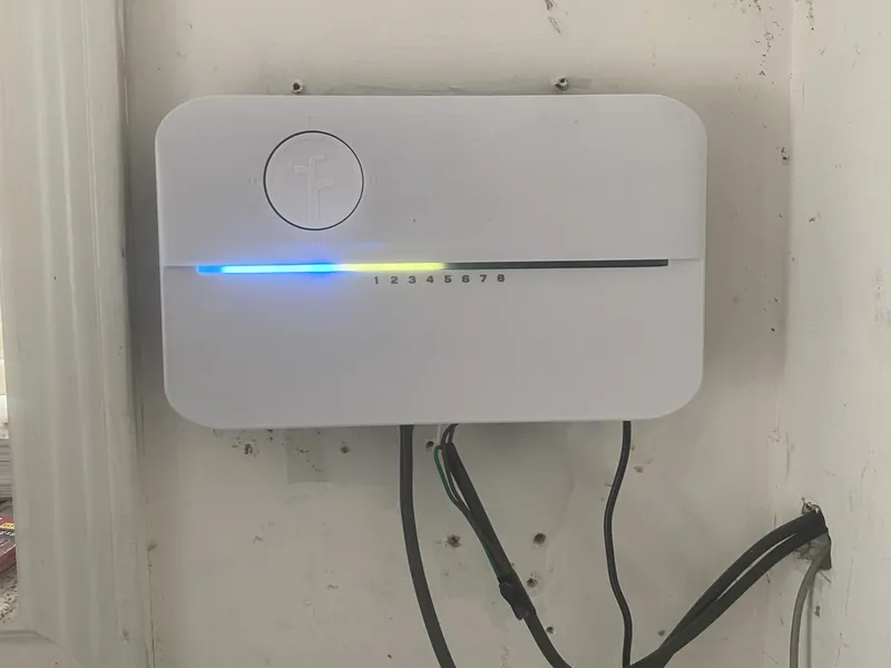 White eight-zone sprinkler controller with a lit blue and yellow LED light bar, mounted on a wall with wires running out the bottom