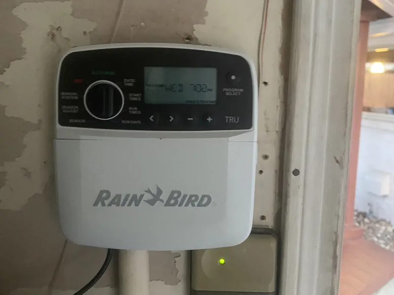 Rain Bird TRU sprinkler controller showing the day and time on its display, mounted on a wall beside a doorway