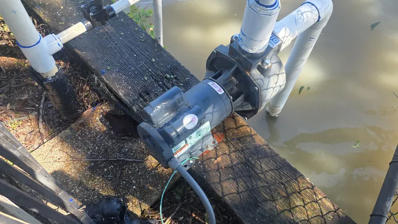 Irrigation pump with white PVC pipes on a wooden edge beside water, behind a chain-link fence