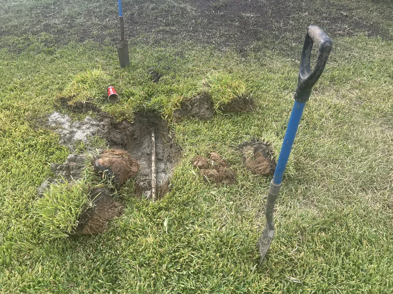 Lawn with sod cut back around a muddy hole over a buried pipe, with a blue-handled shovel standing in the grass