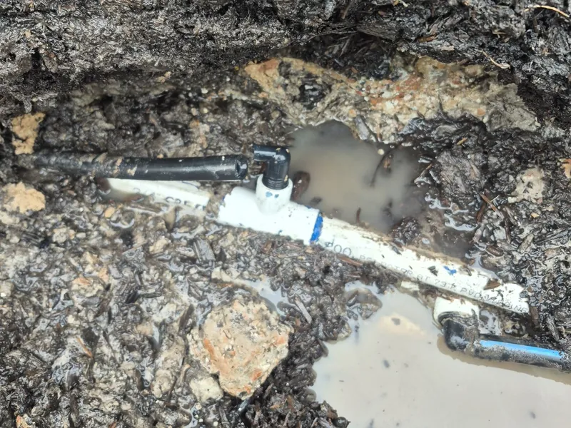 White PVC pipe with blue fittings in a muddy, water-filled trench