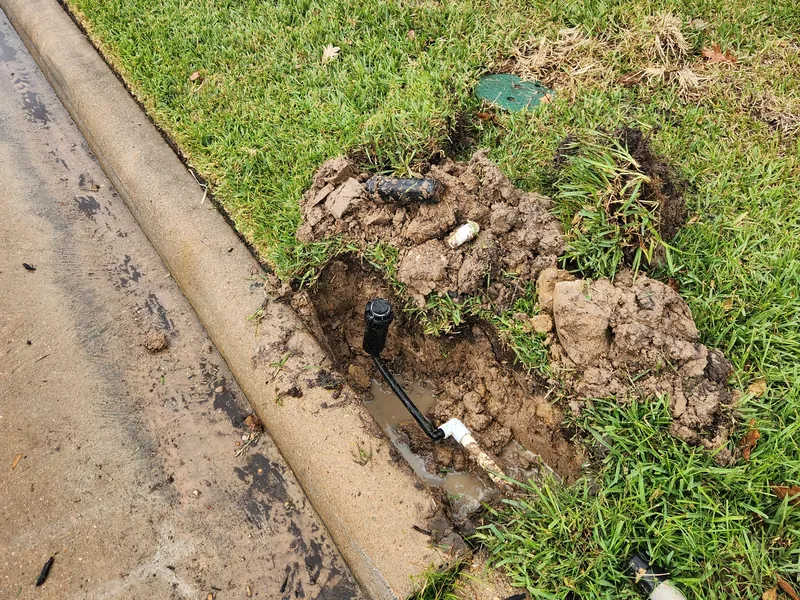 Hole dug at the edge of a lawn beside a curb exposing a sprinkler riser and pipe, with a cut piece of PVC pipe on the grass