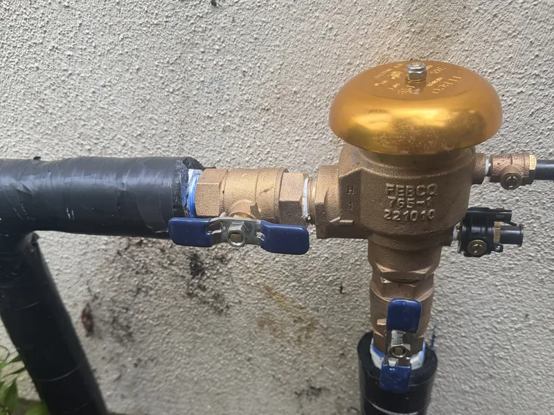Brass pressure vacuum breaker backflow preventer with blue-handled ball valves on insulated pipe against a stucco wall