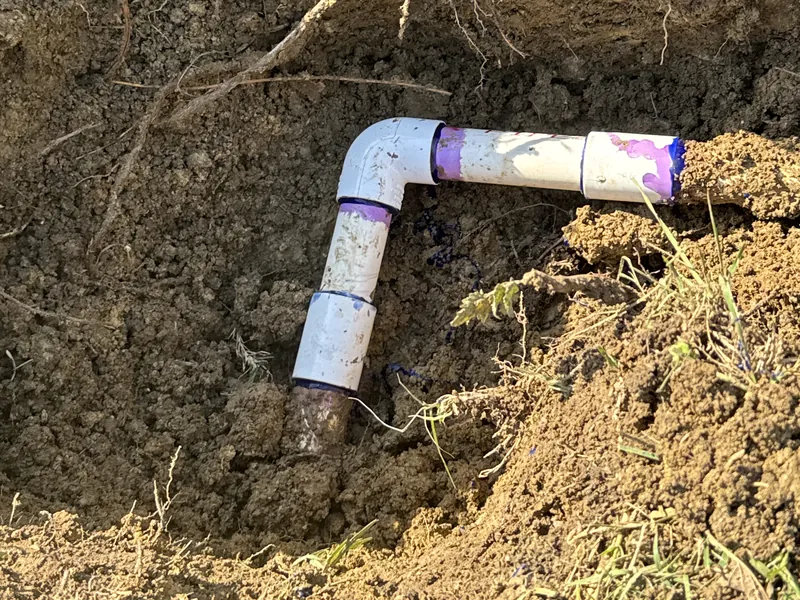 White PVC elbow and couplings with purple primer marks joined to a buried pipe in an open hole