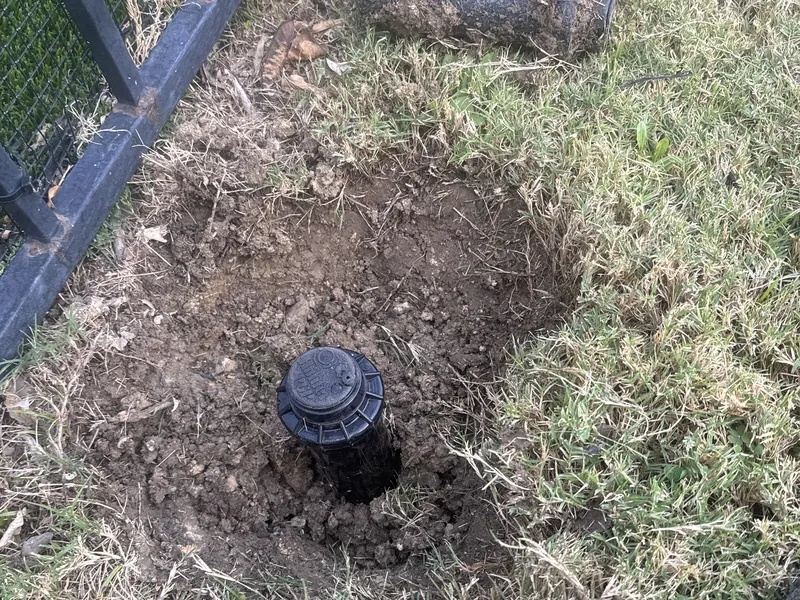 Rotor sprinkler head set in freshly dug soil in a lawn beside a metal fence