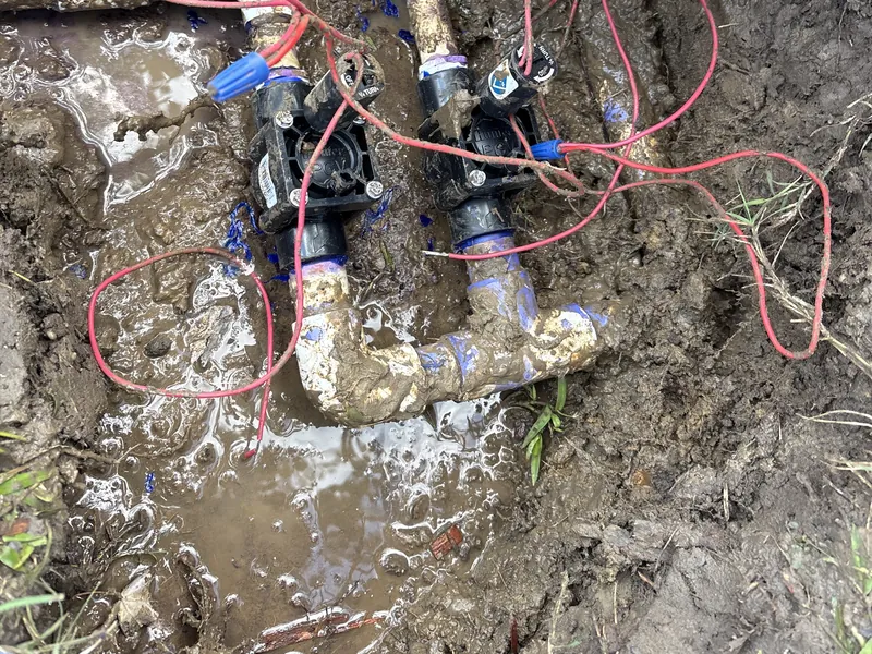 Two sprinkler valves on a muddy PVC manifold in an open hole, with red wires and blue wire connectors