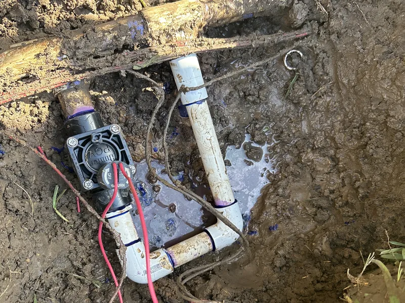Sprinkler valve with red wires connected to white PVC fittings in a dug-out hole